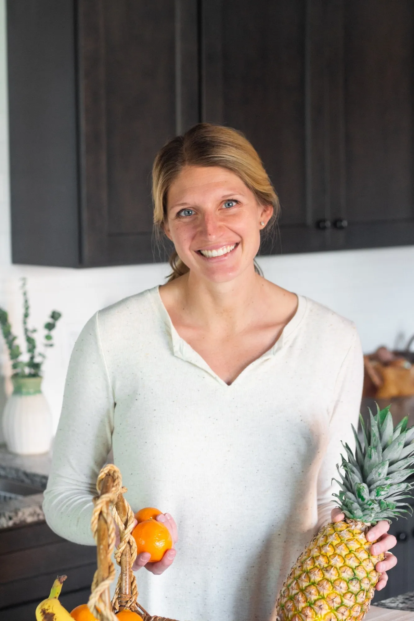 Kelsey Shaughnessy, RDN, holding oranges and a pineapple beside a basket of fruit in a kitchen