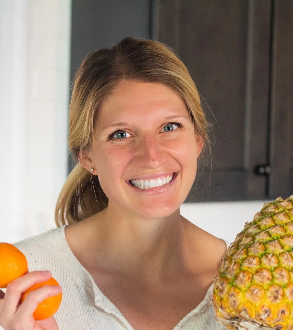 Kelsey Shaughnessy, RDN, smiling and holding oranges and a pineapple in a kitchen